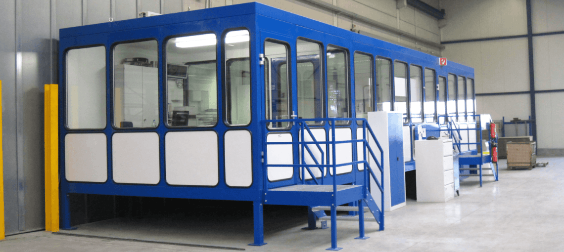Prefab kantoorunit met blauwe staalconstructie en ramen, geplaatst in een industriële hal.