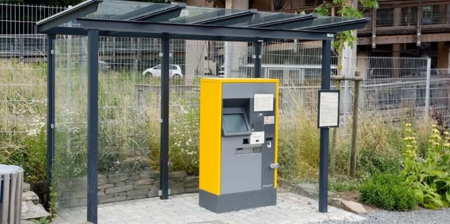 Glazen betaalautomaat overkapping geplaatst over een gele en grijze betaalautomaat op een bestrate ondergrond bij een parkeerterrein.