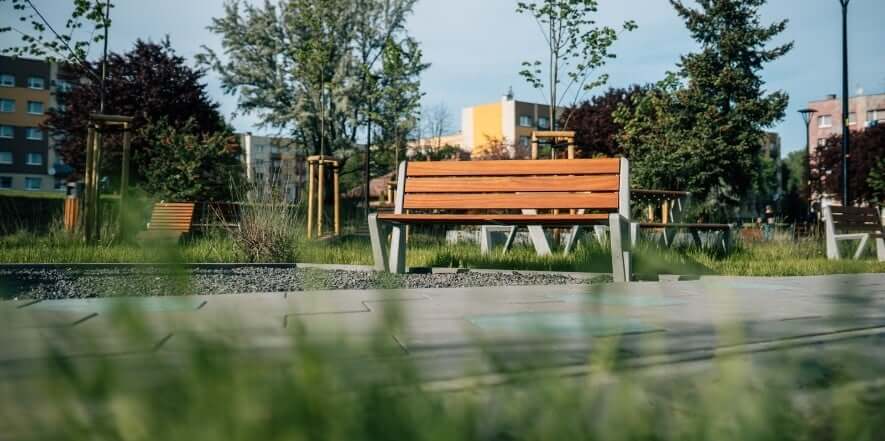 Duurzame houten zitbank met metalen onderstel geplaatst in een groene stadsomgeving met gras, bomen en appartementen op de achtergrond.
