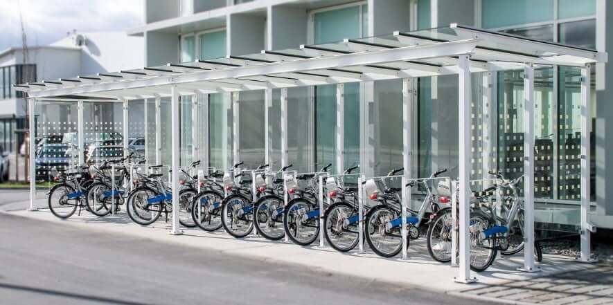 Overdekte fietsenstalling met witte stalen constructie en glazen zijwanden, voorzien van fietsaanleunbeugels op een modern bedrijventerrein.