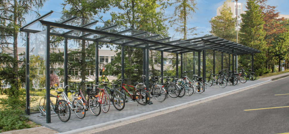 Lange fietsoverkapping van verzinkt staal en glas met fietsenrekken, geplaatst langs een weg in een groene omgeving.