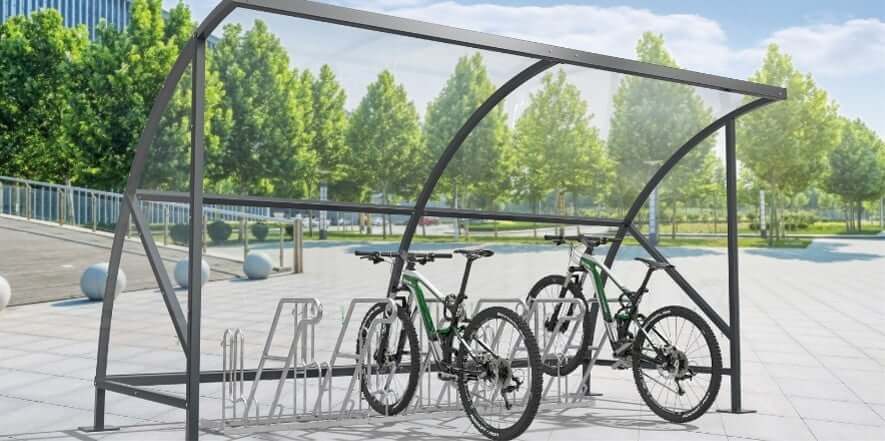 Twee fietsen geparkeerd in een metalen fietsenrek onder een gebogen bouwpakket fietsoverkapping met transparant dak en zijpanelen op een open plein met veel groen op de achtergrond.