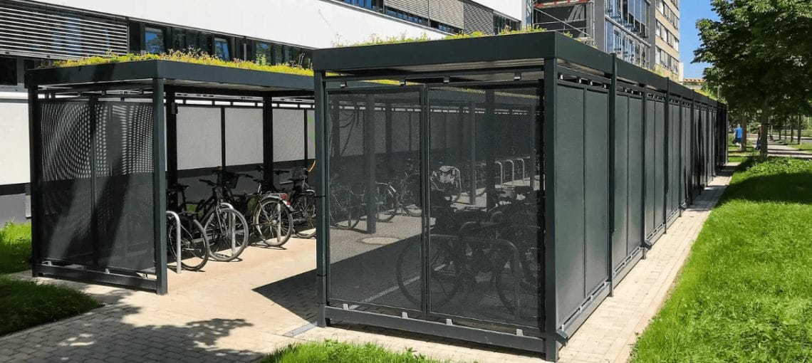 Fietsoverkapping met zwart stalen frame en sedumdak, voorzien van afsluitbare toegang en meerdere fietsenrekken op een betegeld pad naast een kantoorpand.