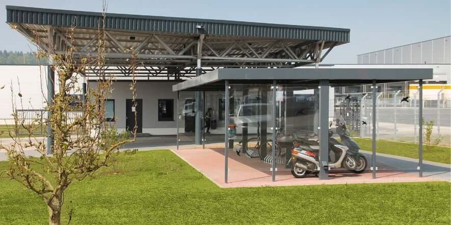 Glazen fietsoverkapping met rijwielrekken en scooters eronder, geplaatst op een terrein met gras en industriële gebouwen