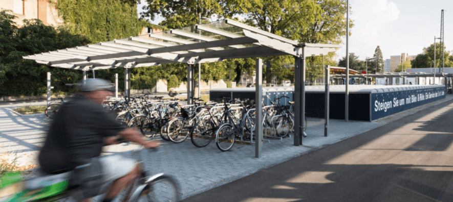 Fiets- en rookoverkapping in één, met een stalen constructie en transparant dak, geplaatst op een betegeld plein. Onder de overkapping staan tientallen fietsen geparkeerd, terwijl op de voorgrond een fietser passeert.