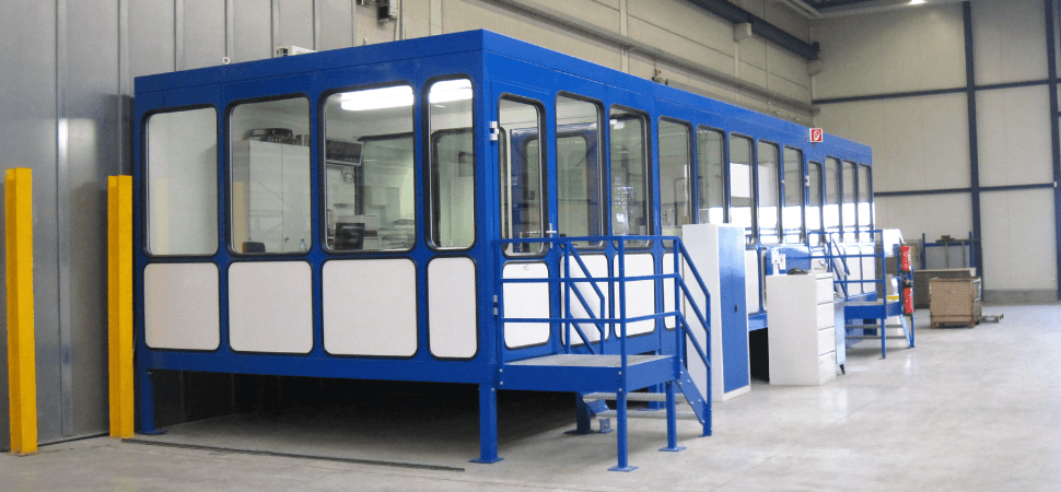 Blauwe maatwerk kantoorunit op verhoogd platform met ramen en trap, geplaatst in een industriële werkomgeving.