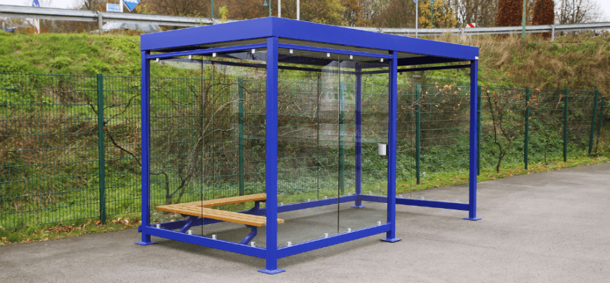 Rookabri voor buiten met blauw stalen frame, transparante glazen wanden en een houten zitbank. De overkapping staat op een verhard terrein langs een groen talud en hekwerk.