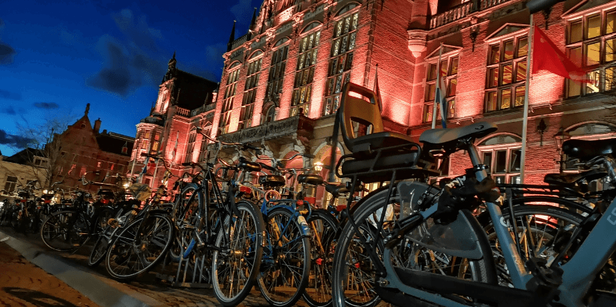 Grote hoeveelheid geparkeerde fietsen voor het verlichte hoofdgebouw van de Rijksuniversiteit Groningen tijdens de avonduren. De historische gevel is rood aangelicht en vormt een contrast met de donkere lucht.