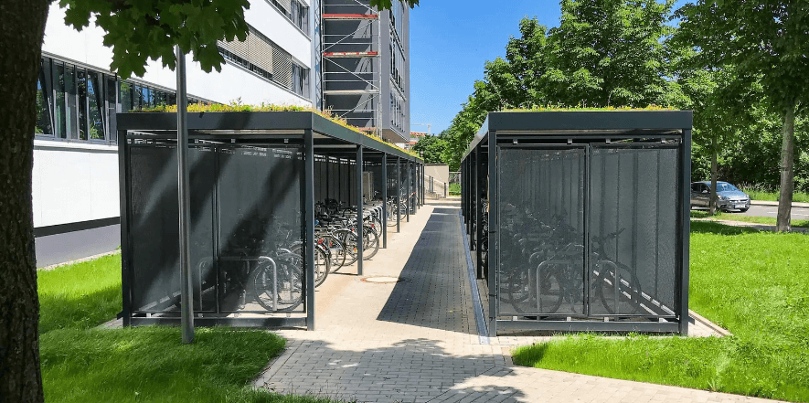 Twee zwarte fietsoverkappingen met sedumdak en geperforeerde zijpanelen, geplaatst aan weerszijden van een pad op een bedrijventerrein. Binnenin staan meerdere fietsen in vaste rekken, met veel groen rondom.
