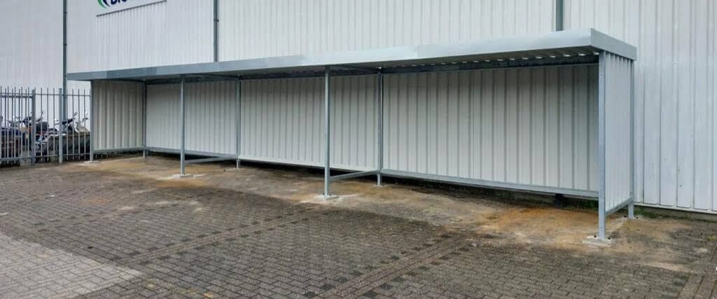 Lange verzinkte fietsoverkapping tegen een gevel, geschikt voor fietsen of materiaalopslag, met een open voorkant en een robuuste stalen constructie.