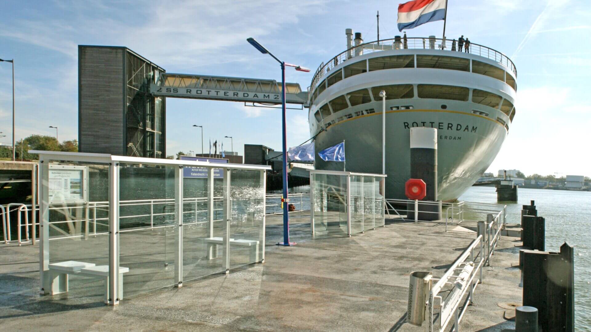 Rookoverkappingen van glas en staal op een kade naast het cruiseschip ss Rotterdam met Nederlandse vlag, bij helder weer. 
