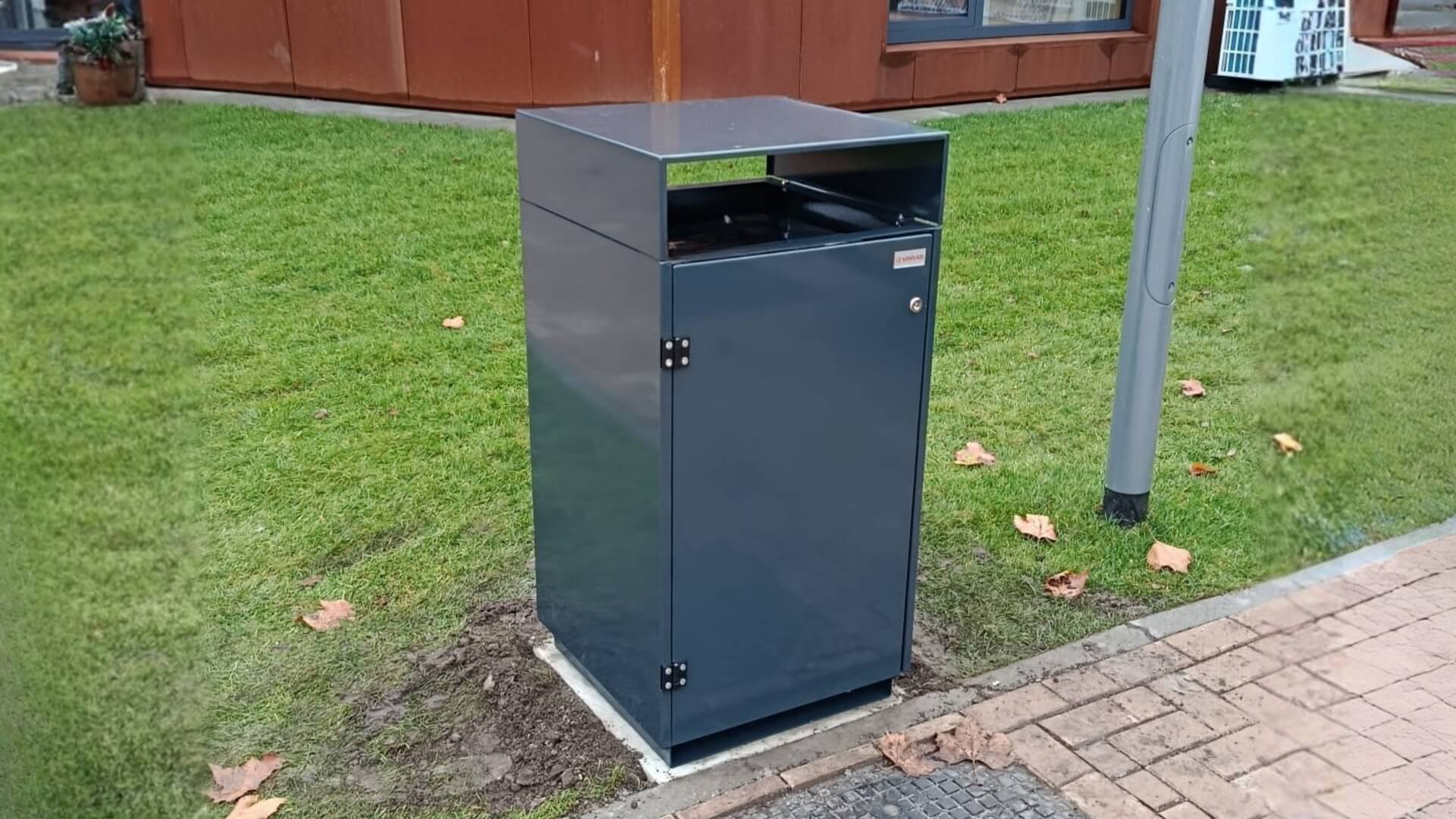 Donkergrijze afvalbak voor buiten van verzinkt staal in RAL 7016, geplaatst op een betonnen voet langs een grasveld bij school in Nijverdal.