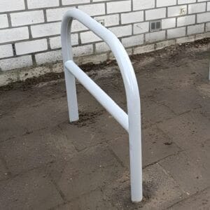 Close-up van een fietsnietje van verzinkt staal, gepoedercoat in RAL-9006 (blank aluminium), stevig in beton gegoten. Het nietje is geplaatst op een betegeld oppervlak voor een witte bakstenen gevel.