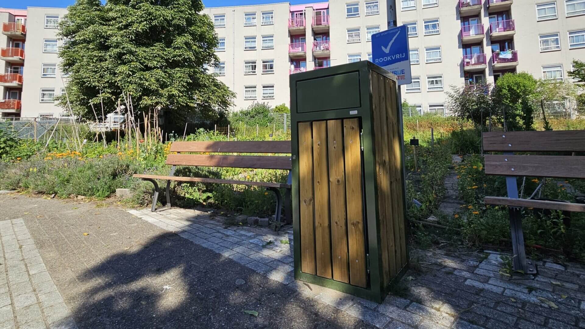 Groene afvalbak van 120 liter met houten latten, geplaatst tussen banken in een tuinachtige omgeving bij een woongebouw.