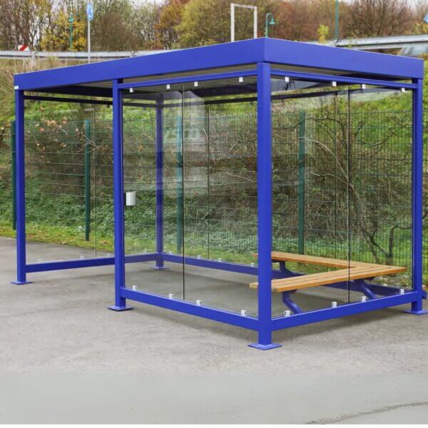 Blauwe Rookabri direct kopen Vrijstaande blauwe rookabri van staal met glazen wanden en houten zitbank, geplaatst op een buitenterrein.