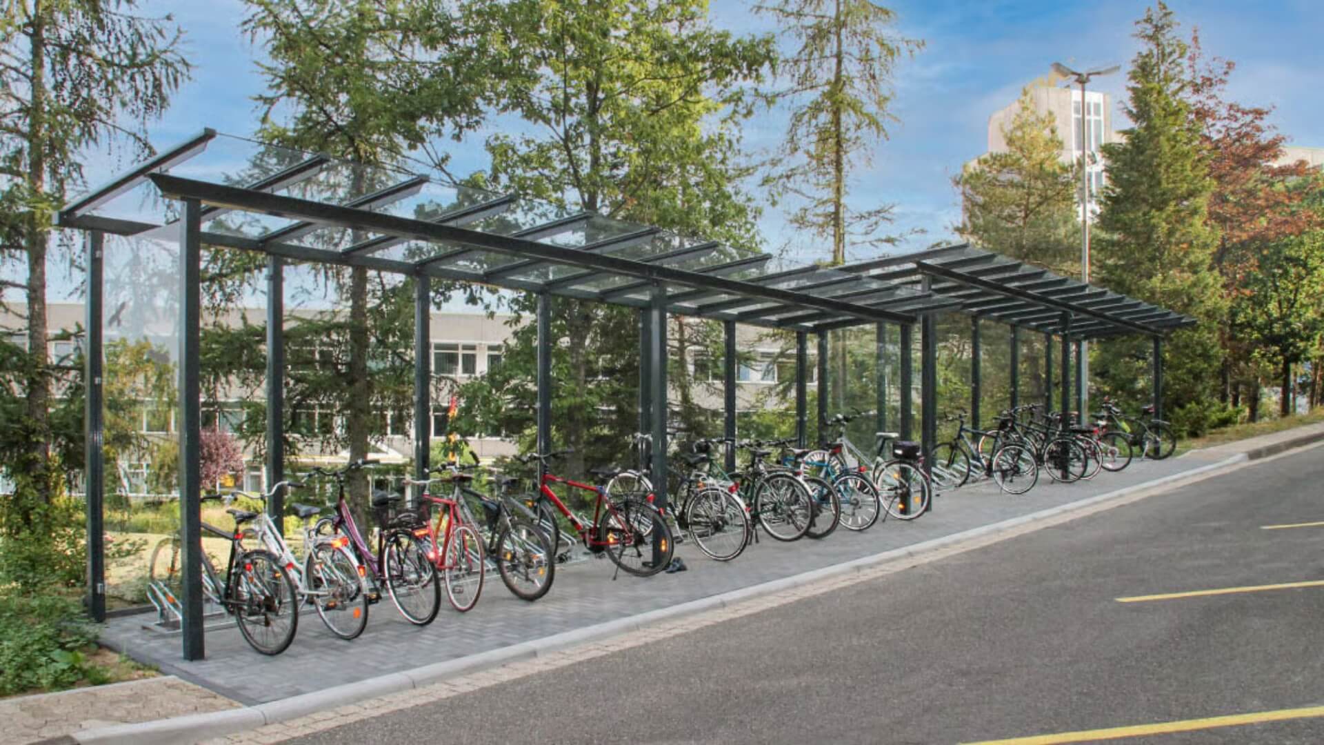 Lange bouwpakket fietsenstalling van glas en staal met meerdere fietsenrekken, geplaatst naast een weg in een groene omgeving.