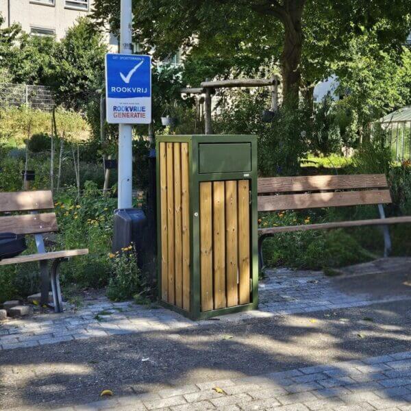 Buiten afvalbak 120 liter online bestellen Buiten afvalbak 120 liter met houten afwerking en RAL 6003 Olijfgroen, geplaatst in een park.