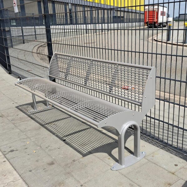 Buitenbank van staal in grijs direct online bestellen Grijze stalen buitenbank met rugleuning, geplaatst langs een hekwerk op een industrieterrein.