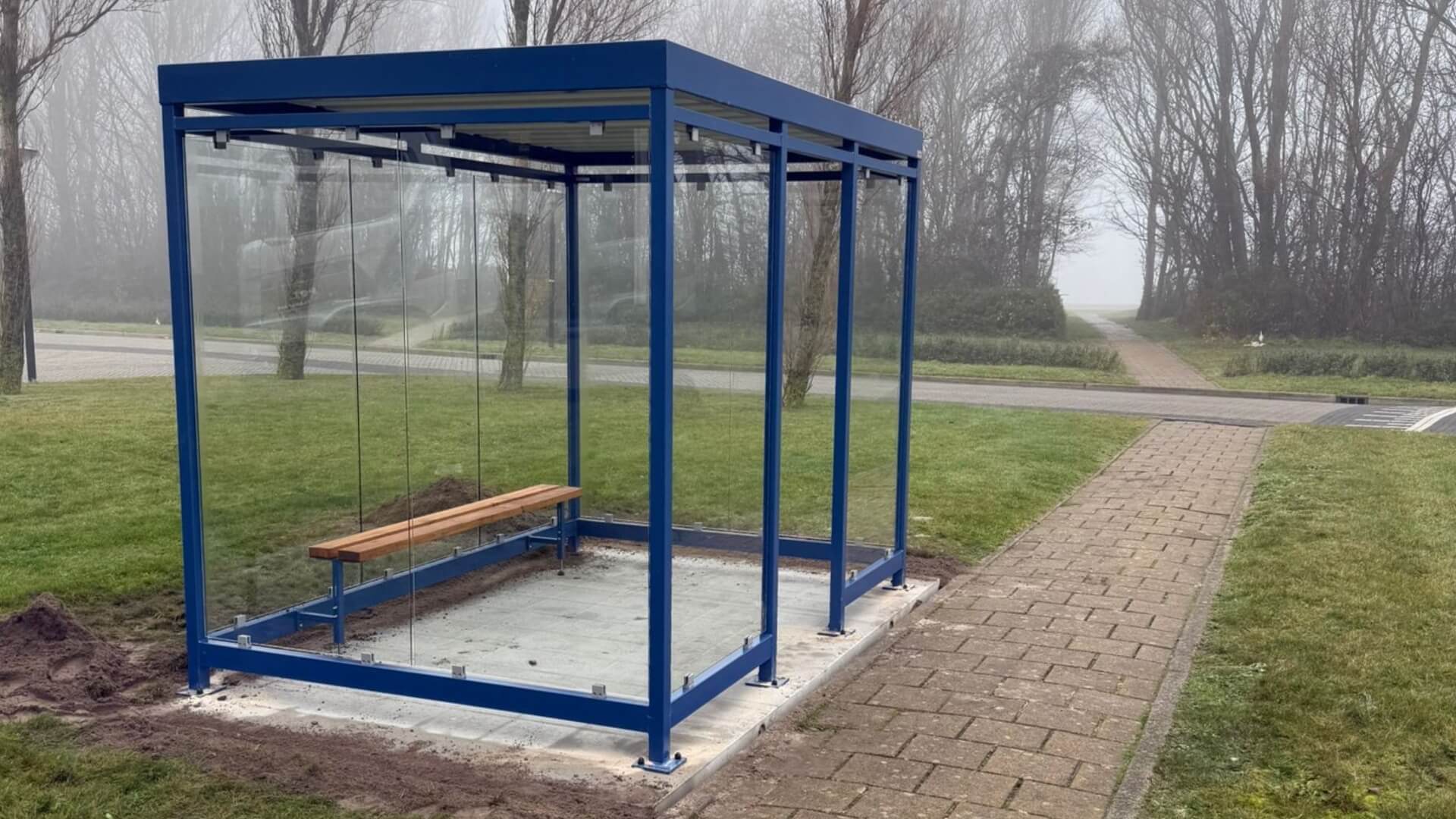 Blauwe rookabri van 3x2 meter met ESG-veiligheidsglas en houten zitbank, geplaatst op een betontegelplatform naast een wandelpad op het terrein van COA Den Helder, gefotografeerd op een mistige ochtend met bomen op de achtergrond.