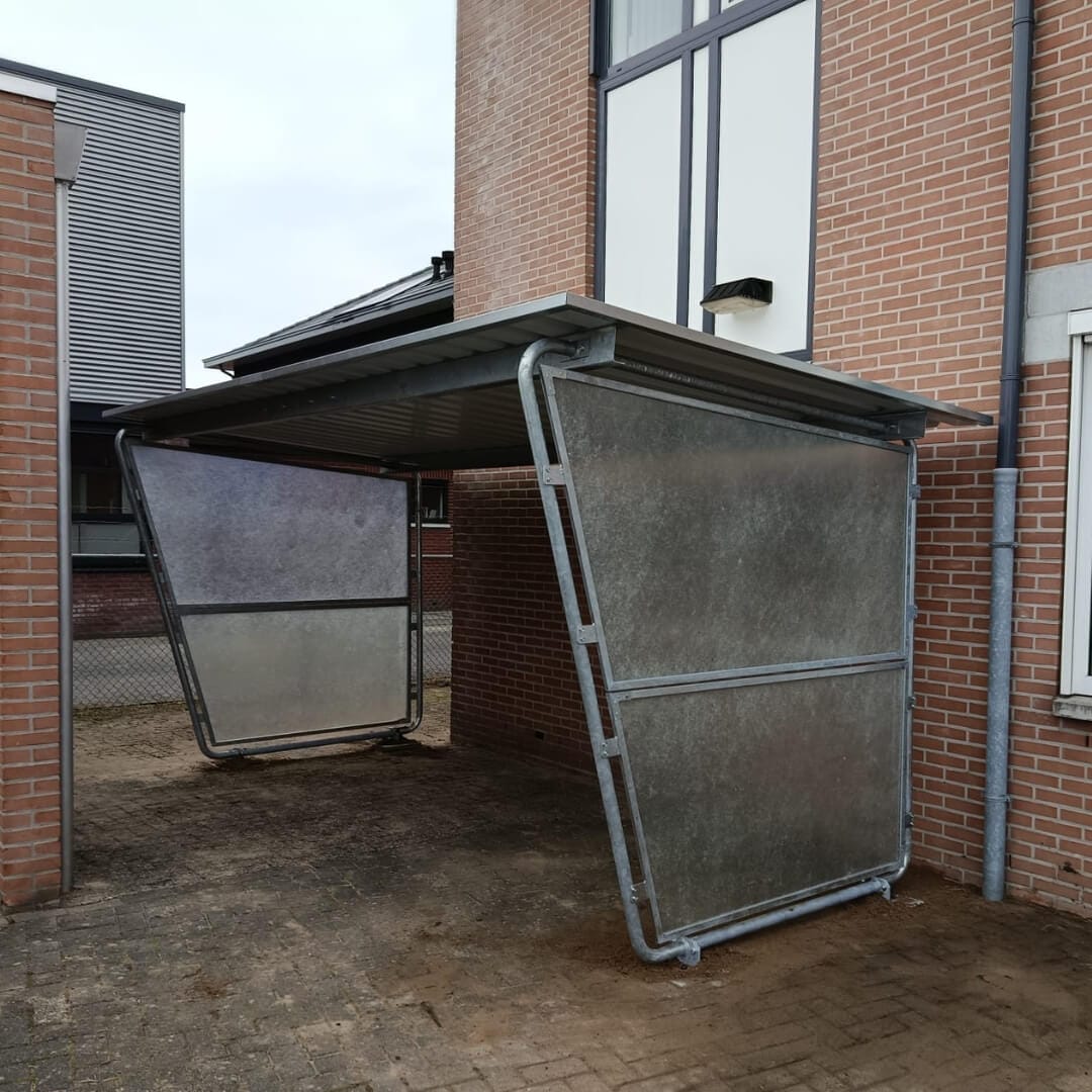Bouwpakket fietsoverkapping met schuin geplaatste metalen zijpanelen en golfplaten dak, gemonteerd tegen een bakstenen muur.
