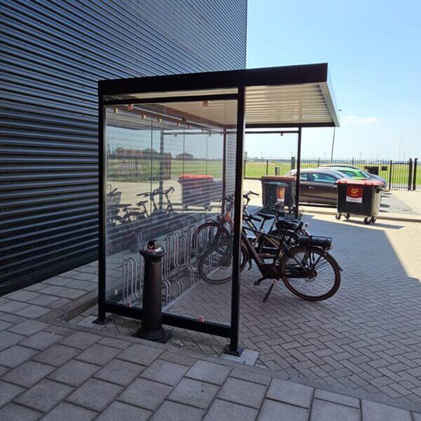 Fietsenstalling voor bedrijven kopen Fietsenstalling met glazen wanden en metalen dak, geplaatst naast een bedrijfspand. Binnenin staan fietsen veilig in een fietsenrek, beschermd tegen regen en wind.