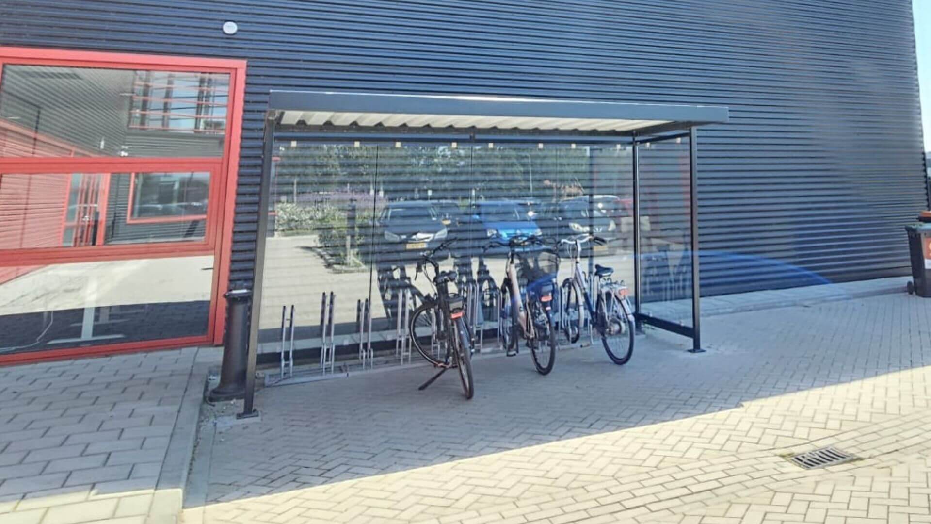 Fietsoverkapping met glazen wanden en metalen frame, geplaatst op een bedrijventerrein. Onder de overkapping staan meerdere fietsen in fietsenrekken.