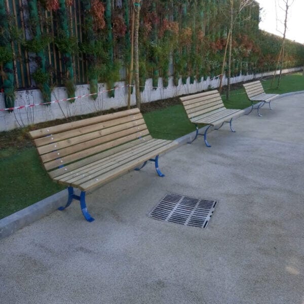 Parkbank van hout direct online bestellen Rij van houten parkbanken met blauwe metalen poten, geplaatst langs een betonnen wandelpad met groenstrook en jonge bomen op de achtergrond.
