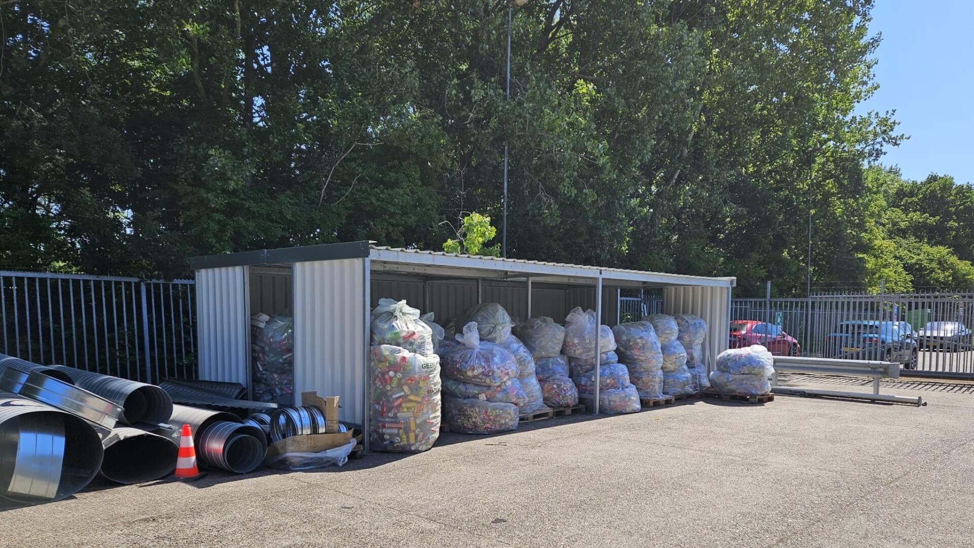 Materiaaloverkapping met zij- en achterwand, gebruikt voor opslag van zakken en metalen onderdelen op een buitenterrein.