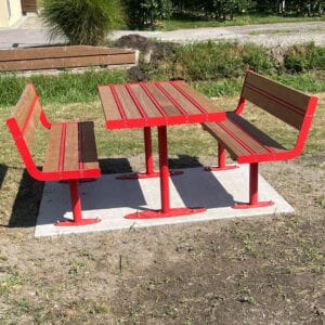 Picknickset met rood stalen frame en houten zitvlakken en tafelblad, geplaatst op een betonnen ondergrond in een groene buitenruimte.