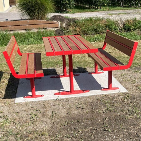 Picknickset met rood stalen frame en houten zitvlakken en tafelblad, geplaatst op een betonnen ondergrond in een groene buitenruimte.