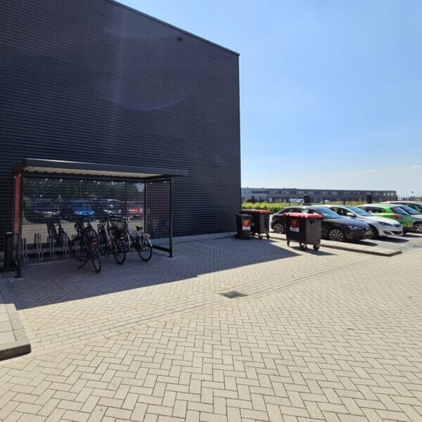 Rijwieloverkapping voor bedrijven aanschaffen Fietsoverkapping met glazen zijpanelen en metalen dak, geplaatst naast een bedrijfsgebouw. Binnenin staan fietsen netjes in een fietsenrek. Op de achtergrond is een parkeerplaats met auto’s en afvalcontainers zichtbaar.