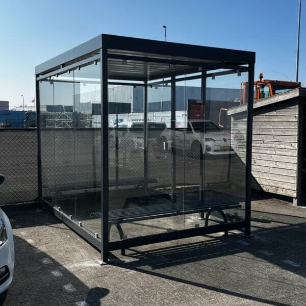 Rokersoverkapping van staal in RAL 7016 met glazen wanden, voorzien van RVS asbak en twee parkbankjes zonder rugleuning op een bedrijventerrein.