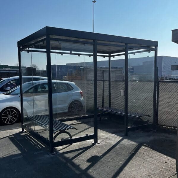 Buiten rokersruimte van staal in RAL 7016 met glazen wanden, RVS asbak en twee bankjes zonder rugleuning op een bedrijventerrein.
