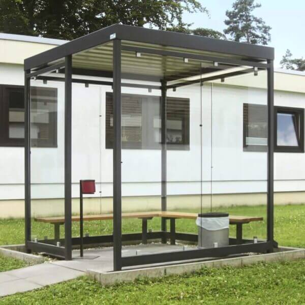 Rokersruimte van 2x2 meter aanschaffen Vrijstaande rokersruimte van 2 bij 2 meter met glazen wanden, zwart stalen frame, zitbank en prullenbak op een grasveld.