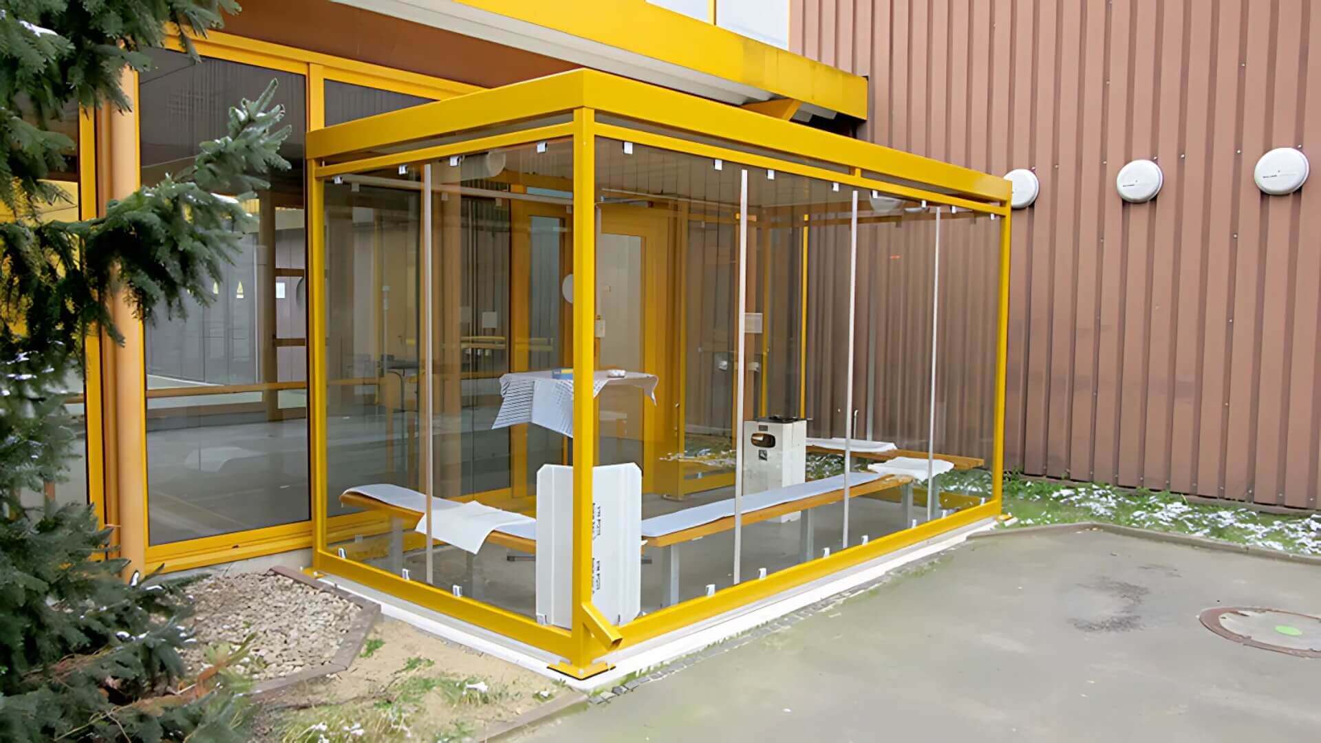 Gele rookoverkapping Keulen van staal en glas, geplaatst tegen een gevel en voorzien van zitbanken voor een comfortabele rookruimte buiten.