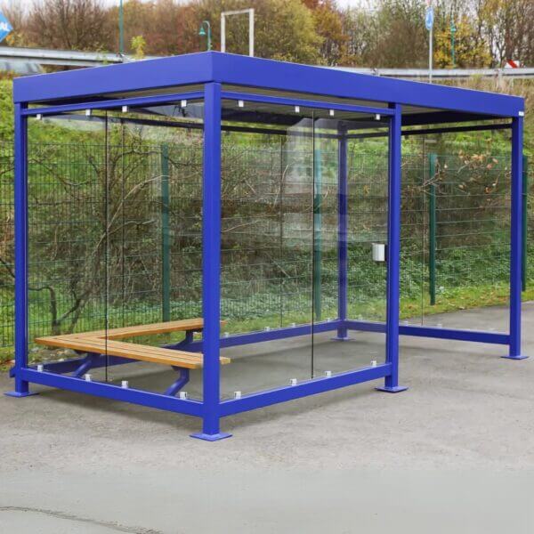 Rookhok in blauw direct online bestellen Blauw stalen rookhok met glazen wanden en houten zitbank, geplaatst op een buitenterrein langs een hekwerk.