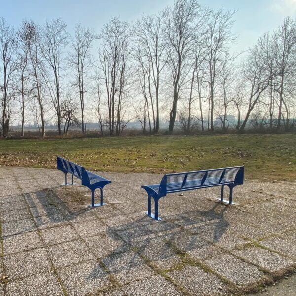 Metalen Twee moderne blauwe metalen parkbanken met rugleuning op een betegeld plein met uitzicht op een grasveld en kale bomen in de achtergrond.