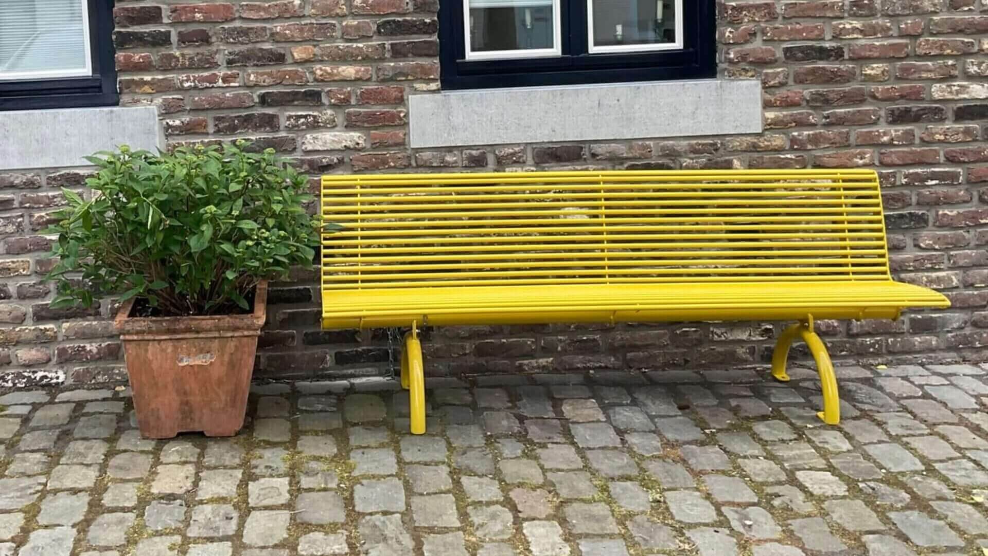 Parkbank van staal in het geel met een lengte van 198 cm tegen een bakstenen muur