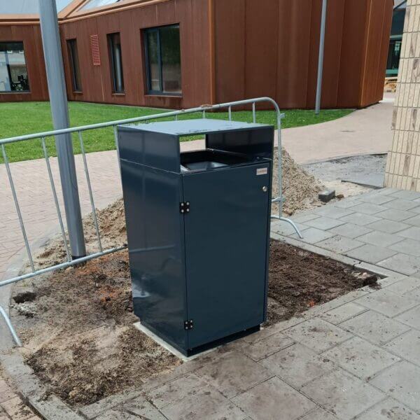 Afvalbak buiten laten kopen voor schoolgebouwen Donkergrijze afvalbak van verzinkt staal met boveninworp, geplaatst op een net aangelegd trottoir bij school Reggesteyn in Nijverdal. De afvalbak is afgewerkt in kleur RAL 7016 en omringd door tijdelijke hekwerken.