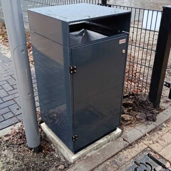 Afvalbak buiten aanschaffen voor entree of schoolplein Buitenafvalbak van verzinkt staal in RAL 7016, geplaatst bij een hekwerk en lantaarnpaal op het terrein van Reggesteyn in Nijverdal. De afvalbak is voorzien van een boveninworp en stevig gemonteerd op een betonnen voet.