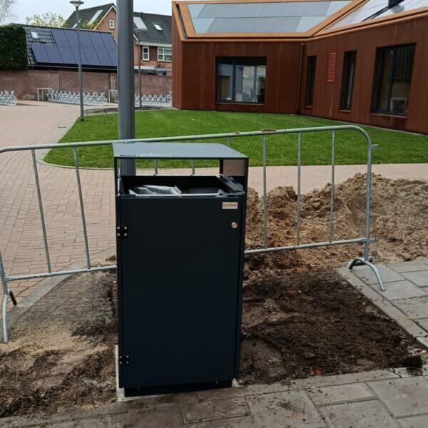 Afvalbak buiten kopen voor scholen en instellingen Donkergrijze buitenafvalbak in RAL 7016 van verzinkt staal, geplaatst voor een modern schoolgebouw bij Reggesteyn in Nijverdal. De afvalbak staat naast een bouwhek en is nog omringd door verse grond.