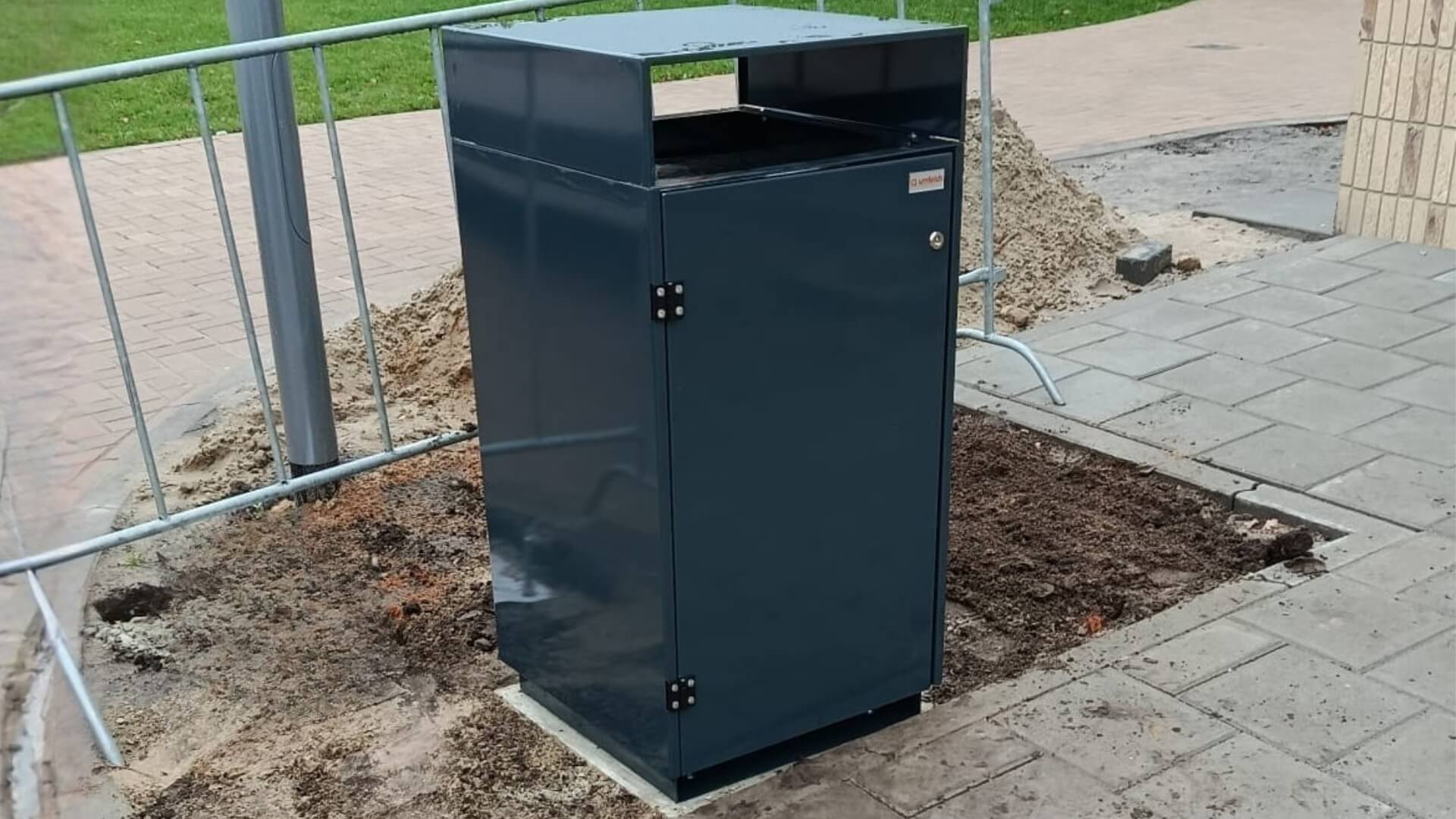 Buitenafvalbak van verzinkt staal met boveninworp, geplaatst op een betonnen fundering langs het trottoir bij school Reggesteyn in Nijverdal. De bak is afgewerkt in RAL 7016 en staat naast tijdelijke hekwerken en bestrating.