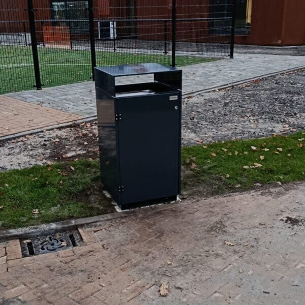 Buitenafvalbak eenvoudig online bestellen Buitenafvalbak van verzinkt staal in RAL 7016, geplaatst aan de rand van een betegeld looppad bij school Reggesteyn in Nijverdal. Op de achtergrond zijn een sportveld en een schoolgebouw zichtbaar.