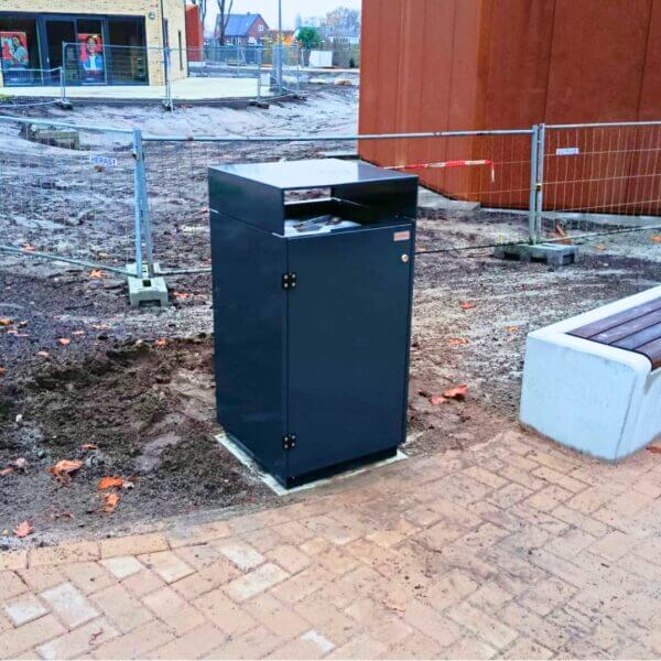 Buitenafvalbak kopen voor schoolpleinen Donkergrijze buitenafvalbak van verzinkt staal met moderne, strakke vormgeving op het terrein van school Reggesteyn in Nijverdal. De afvalbak is uitgevoerd in kleur RAL 7016 en stevig verankerd naast een betonnen zitbank.