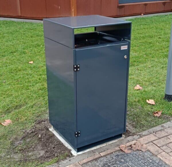 Donkergrijze buitenafvalbak van verzinkt staal met boveninworp, geplaatst op een betonnen voet langs een grasveld bij Reggesteyn in Nijverdal. De bak is afgewerkt in RAL 7016 en staat bij een modern schoolgebouw.