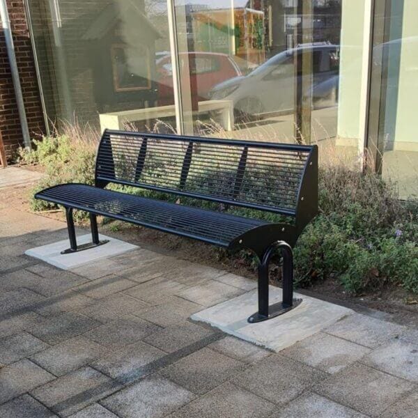 Stalen buitenbank Carinzia kopen voor jouw buitenruimte Stalen buitenbank Carinzia met poedercoating geplaatst op verankerde voetplaten naast een gevel met beplanting.