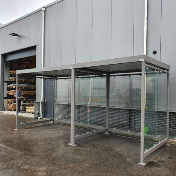 Dubbele rookabri van elk 2x2 meter, uitgevoerd in grijs, verankerd op asfalt en geplaatst bij een bedrijfsgebouw.