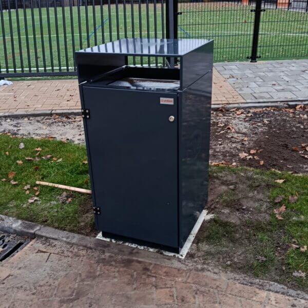 Duurzame afvalbak buiten aanschaffen voor schoolterreinen Donkergrijze buitenafvalbak in RAL 7016 van verzinkt staal, geplaatst bij een sportveld op het terrein van Reggesteyn in Nijverdal. De robuuste afvalbak staat stevig gemonteerd langs het trottoir.