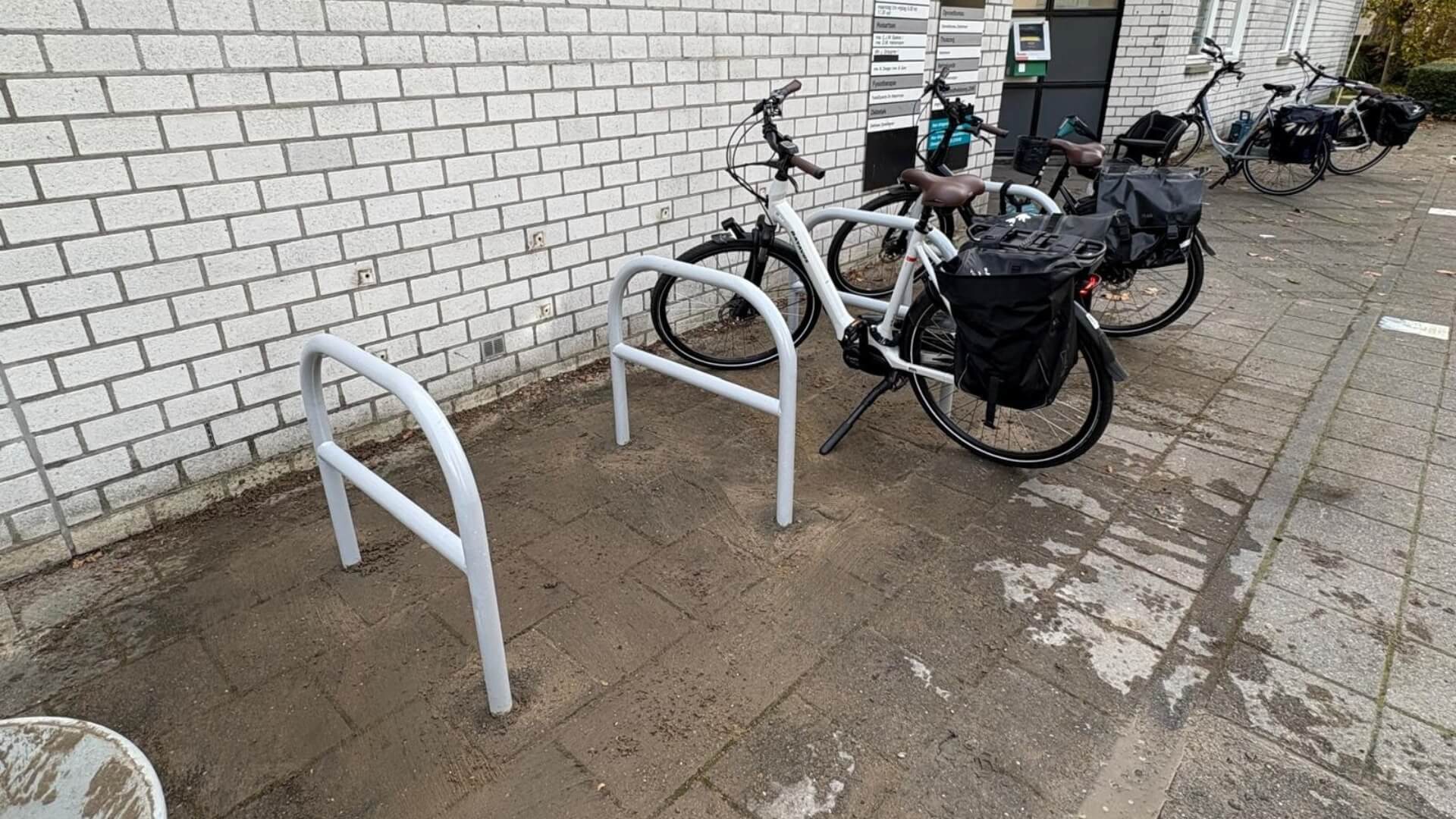 Fietsnietjes van verzinkt staal met tussenbuis, gepoedercoat in RAL-9006, geïnstalleerd bij Gezondheidscentrum de Watertoren in Zoetermeer. Enkele fietsen staan geparkeerd, terwijl andere beugels nog vrij zijn op het natte bestrate plein.