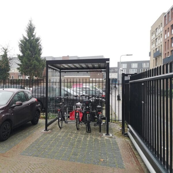 Moderne zwarte fietsoverkapping van staal met ESG glazen zijwanden, geplaatst op betonpoeren en voorzien van fietsrekken voor veilige stalling van 4 tot 6 fietsen.